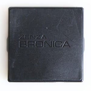 Op de afbeelding: Een zwart vierkant camera-accessoire met de in reliëf aangebrachte tekst "ZENZA BRONICA" in het midden aan de bovenkant. Het accessoire heeft een licht gestructureerd oppervlak en afgeronde hoeken. Het item staat tegen een witte achtergrond.