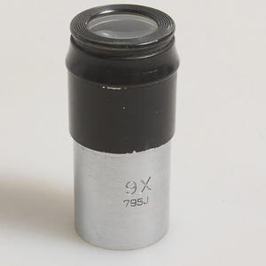 May include: A cylindrical microscope eyepiece with a black top section featuring a glass lens and a silver metal lower barrel. The silver portion is engraved with the text 9X and 795J, indicating the magnification power and model identification.