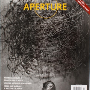 Op de afbeelding: Omslag van het tijdschrift Aperture met het woord "APERTURE" in geel, omcirkeld door een gele ring. De achtergrond is een zwart-wit afbeelding van verwarde draden. Tekst bevat "Roger Ballen's Live Wires" en artikel titels.