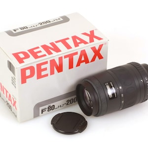 Könnte beinhalten: Ein schwarzes Pentax F80-f4.7-200 Objektiv mit einer schwarzen Objektivkappe. Das Objektiv liegt auf einer weißen Oberfläche. Das Objektiv befindet sich in einer weißen Box mit dem Wort "PENTAX" in Rot.