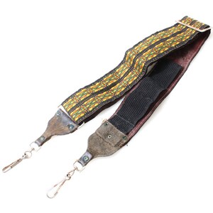 Op de afbeelding: Een vintage cameraband met een bruine leren achterkant en een geweven gele, groene en zwarte band. De band heeft twee metalen clips.