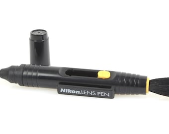 May include: A black Nikon Lens Pen with a yellow button and a black brush on the end. The pen is used to clean camera lenses.