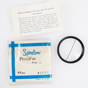 May include: A Spiratone ProxiFar lens filter kit. The kit includes a 49 mm filter, a black lens ring, and a printed instruction sheet. The box is white with blue accents and text. The filter is made in Japan.