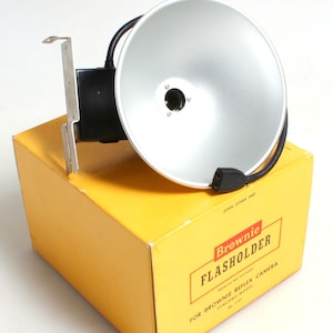 May include: A silver flash holder for a Brownie Reflex camera. The flash holder is attached to a yellow box with the text "Brownie Flasholder for Brownie Reflex Camera Synched Model No. 1133" printed on it.