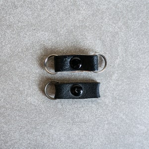 以下が含まれることがあります： シルバーのキーリングが付いた黒い革製のキーホルダー2つ。各キーホルダーの中央には黒いボタンが付いています。キーホルダーは、明るい灰色のテクスチャのある表面に表示されています。