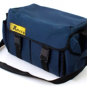 Puede incluir: Una bolsa de cámara de lona azul con una correa de hombro negra y hebillas negras. La bolsa tiene un logotipo amarillo que dice "Amigo".
