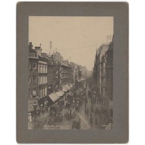 May include: A vintage black and white photograph of a bustling city street scene. The image captures a wide street lined with multi-story buildings, awnings, and horse-drawn carriages. Numerous pedestrians are visible, creating a sense of activity and historical charm.