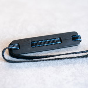 May include: Black camera strap with a blue stripe and a black plastic grip that says "OLYMPUS" in white lettering.