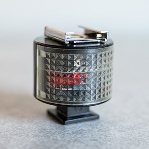 Peut inclure: Un diffuseur de flash noir avec une lumière rouge et un support en métal argenté. Le diffuseur a une grille.