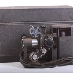 Puede incluir: Un proyector de películas vintage negro con una caja de metal y un carrete de película plateado. El proyector está sentado en una mesa con un cable negro.