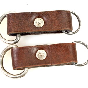以下が含まれることがあります： 銀色の金属製のリングが付いた、茶色の革製のキーホルダーが2つ。キーホルダーにはそれぞれ銀色の金属製のボタンが付いています。