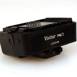 May include: Black Vivitar PM/C flash unit with a lock button and a small arrow pointing to the right. The flash unit has a black plastic body and a silver metal connector.
