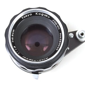 Può includere: Un obiettivo per fotocamera nero con dettagli argentati. L'obiettivo ha la scritta "Tokyo Kogaku Japan No.283040 Auto-Topocor 1:1.8 f=5.8cm" stampata su di esso.