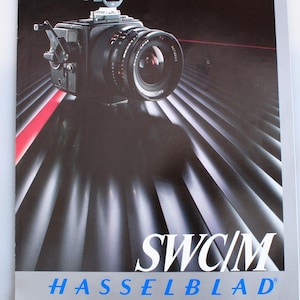 Puede incluir: Una cámara Hasselblad SWC/M negra y plateada con una tapa de lente en el lente. La cámara está sentada sobre una superficie con rayas negras y plateadas. El texto "SWC/M" y "HASSELBLAD" está impreso en blanco sobre un fondo plateado. El texto "SALES & SERVICE" está impreso en negro sobre un fondo blanco. El texto "BERKIE'S PHOTO CENTER" está impreso en negro sobre un fondo blanco. El texto "525 E. Ohio St. Pg., PA 15212-5593" está impreso en negro sobre un fondo blanco. El texto "(412) 231-1718" está impreso en negro sobre un fondo blanco.