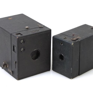 May include: Two vintage black box cameras are displayed side-by-side. The cameras have a rectangular shape with a lens, viewfinder, and various dials. The camera on the left is slightly larger than the one on the right. The cameras appear to be made of leather.
