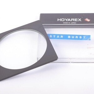 Può includere: Un filtro Hoya Star Burst nero con una cornice quadrata e una lente in vetro trasparente. Il filtro è accanto alla sua confezione originale, che reca l'etichetta "HOYAREX MADE IN JAPAN". La confezione ha anche un'etichetta blu con la scritta "STAR BURST".