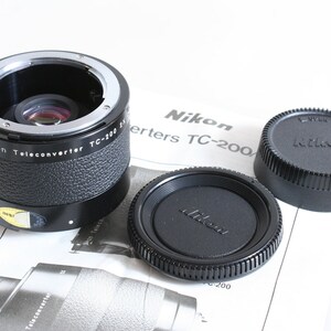 Könnte beinhalten: Ein schwarzes Nikon Telekonverter TC-300 2x Objektiv mit einem silbernen Ring und einer schwarzen Objektivkappe. Das Objektiv liegt auf einer weißen Oberfläche mit einer Nikon Bedienungsanleitung.