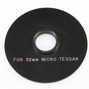 May include: A black circular lens adapter with a central aperture. The text "FOR 32mm MICRO TESSAR" is printed in white. The adapter is designed for use with a 32mm Micro Tessar lens.