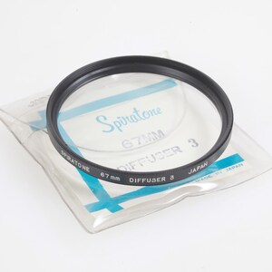 Puede incluir: Un filtro de lente de cámara negro Spiratone 67 mm Diffuser 3. El filtro está en un paquete de plástico transparente con detalles en azul y blanco. El texto "Spiratone", "67 mm" y "Diffuser 3" es visible en el filtro y el embalaje. Hecho en Japón.