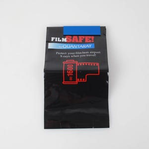 Könnte beinhalten: Schwarze Film Safe-Tasche mit dem Text "FILM SAFE!" und "QUANTARAY". Die Tasche hat einen blauen Streifen oben und eine rote Grafik einer Filmrolle. Auf der Tasche steht "Schützen Sie Ihren Film vor Röntgenstrahlen am Flughafen, wenn Sie reisen!"