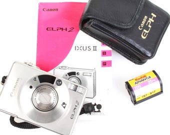 Pode incluir: Uma câmera compacta Canon Elph 2 prateada com uma capa de couro preta e um rolo de filme Kodak Advantix 200. A câmera tem uma lente zoom e um flash embutido.