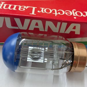 Peut inclure: Une lampe de projecteur Sylvania vintage avec un capuchon bleu et une base couleur cuivre. L'ampoule en verre transparent abrite des composants internes complexes. La boîte rouge derrière la lampe indique "Projector Lamp" et "SYLVANIA" en lettres blanches.