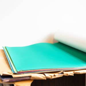 May include: A stack of colorful paper sheets, including a bright teal sheet, on top of a brown cardboard surface. A white roll of paper is partially unrolled and resting on top of the stack.