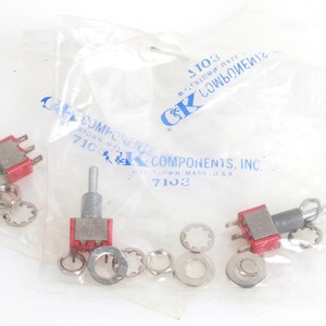 Puede incluir: Varios interruptores de palanca con bases rojas y palancas plateadas son visibles, junto con varias arandelas y sujetadores. Los interruptores están empaquetados en una bolsa de plástico transparente con el texto "C&K COMPONENTS, INC.".