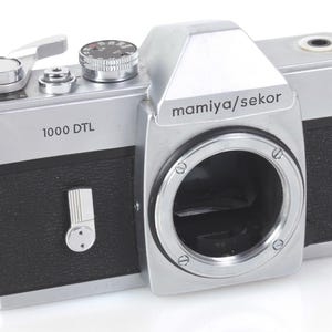 May include: A silver and black Mamiya/sekor 1000 DTL 35mm film camera body. The camera features a silver top plate with a film advance lever, shutter speed dial, and a central pentaprism housing. The body has a black textured grip and metal strap lugs.