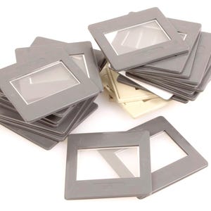 May include: A collection of gray plastic slide mounts with clear windows, stacked and scattered against a white background. Some mounts are stacked in neat piles, while others are scattered. The slides are a vintage item.