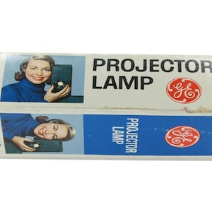 May include: Vintage GE Projector Lamp box. The box is white with blue accents and features a smiling woman holding a projector. The text "PROJECTOR LAMP" is displayed prominently. The GE logo is also visible.