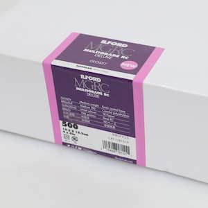 以下が含まれることがあります： 紫とピンクのラベルが付いた白い箱。ラベルには「ILFORD MGRC MULTIGRADE RC DELUXE GLOSSY」と「NEW」と書かれています。箱には16.3 x 21.5 cmのフォトペーパーが500枚入っています。