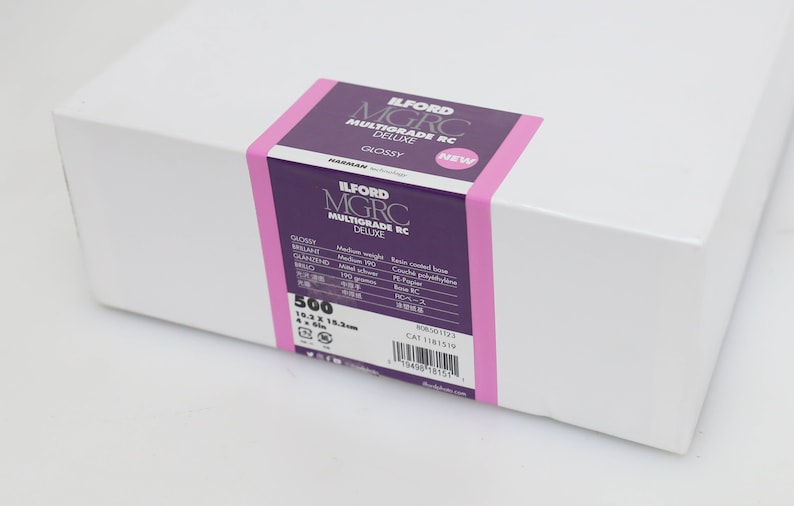 以下が含まれることがあります： 紫とピンクのラベルが付いた白い箱。ラベルには「ILFORD MGRC MULTIGRADE RC DELUXE GLOSSY」と「NEW」と書かれています。箱には16.3 x 21.5 cmのフォトペーパーが500枚入っています。