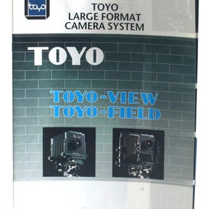 May include: A black and white brochure for Toyo Large Format Camera System. The brochure features the text "TOYO" in large blue letters and two images of Toyo-View and Toyo-Field cameras. The brochure also includes the text "SAKAI SPECIAL CAMERA MFG.CO.,LTD."