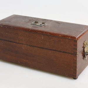 Puede incluir: Una caja rectangular de madera con tapa con bisagras y cierre metálico. La caja es de color marrón oscuro con vetas de madera visibles y un pestillo y cerradura de color latón. La caja tiene patas pequeñas.