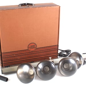 Peut inclure: Une mallette de transport en carton marron avec une poignée noire et le texte "Foto-Lite" dans un logo circulaire, accompagnée d'une barre lumineuse en métal argenté avec quatre grands abat-jours sphériques gris métallisé et un petit outil en plastique noir.
