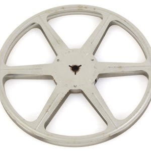 Puede incluir: Una bobina de película vintage de color gris claro. La bobina tiene un diseño de estrella con seis radios que irradian desde un cubo central. El cubo tiene una serie de pequeños agujeros. La bobina es de metal y parece usada.