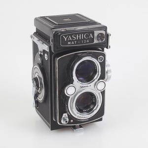 May include: A black Yashica Mat-124 twin-lens reflex camera. The camera has two lenses, a silver lens ring, and the text "Yashica Mat-124" on the top. The camera has a silver winding knob on the side and a silver lens ring.