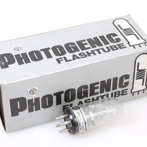 以下が含まれることがあります： 「PHOTOGENIC FLASHTUBE」の黒い文字が入った銀色の箱。黒いベースと金属ピンが付いた透明なガラス製フラッシュチューブが箱の前にあります。箱にはフラッシュチューブのグラフィックがあります。