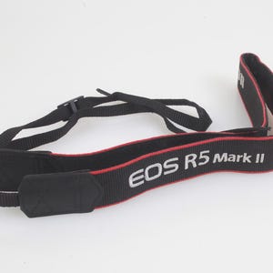 May include: A black camera neck strap featuring red piping along the edges and the white text EOS R5 Mark II embroidered on the front. The strap includes adjustable black nylon webbing and reinforced black fabric ends for secure camera attachment.