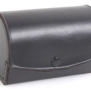 May include: A black leather case with a rounded top and a rectangular base. The case has a button closure and a stitched seam. The interior edges are red. The case is likely for storing a camera or other small items.