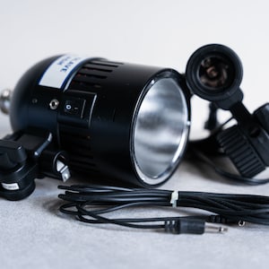 May include: A black studio light with a silver reflector, a power cord, and a light bulb socket. The light has a switch on the side and a clamp for attaching it to a stand.