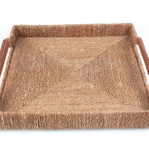 May include: A square woven seagrass tray with a natural tan color and a textured, braided pattern. The tray features two integrated side handles wrapped in a smooth, brown faux leather material, providing a contrast to the woven base structure.