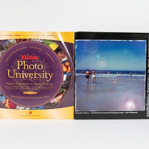 May include: Two books about photography. The book on the left is purple with a yellow border and says "Kodak Photo University" with the text "Higher Education in Picture Taking". The book on the right is black with a colorful image of a beach scene and says "Polaroid Studio 3" with the text "An Interactive Guide for Professional Photographers".