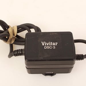 May include: A black Vivitar DSC-3 device with a black cord and a tan rubber band. The device has the text "Vivitar DSC-3" printed on it. The cord is looped and connected to the device.