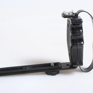 May include: Black camera flash bracket with a handle and a mounting plate. The bracket has a shoe mount for attaching a flash unit. The handle is a black strap. The mounting plate has a screw for attaching to a camera.