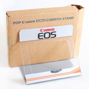 May include: A cardboard box labeled "POP Canon EOS CAMERA STAND" with a clear plastic display stand in front. The Canon logo is red, and the EOS text is black.