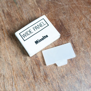 Peut inclure: Une boîte blanche avec le texte "WIDE PANEL" et "Minolta" imprimé dessus. Un filtre en plastique blanc est à côté de la boîte.