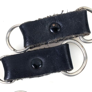 以下が含まれることがあります： 銀色の金属製のリングが付いた黒い革製のキーホルダーが2つ。キーホルダーにはそれぞれ黒いボタンが付いています。