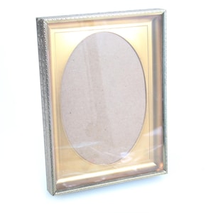 May include: A rectangular picture frame with an oval opening. The frame has a silver-colored, textured border and a gold-colored interior. The oval opening is covered with clear plastic.